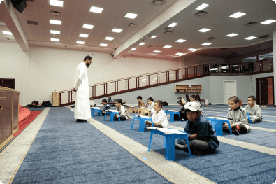 Teacher walking among students reading the Quran in the prayer hall at Abu Huraira Center